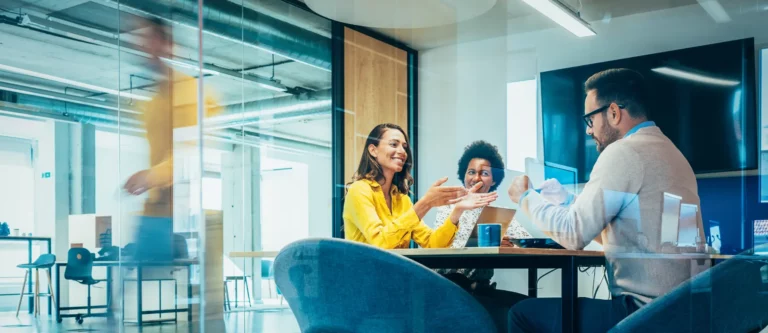 Three people sit around a table in a modern office, having a discussion about workforce strategy and smiling. One woman gestures while talking, another woman listens, and a man faces them. A blurred figure walks by outside the glass meeting room.