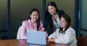 Three women sit around a table, smiling and looking at a laptop screen. One stands behind, pointing at the laptop as they discuss a cost-efficient growth strategy displayed on the screen.