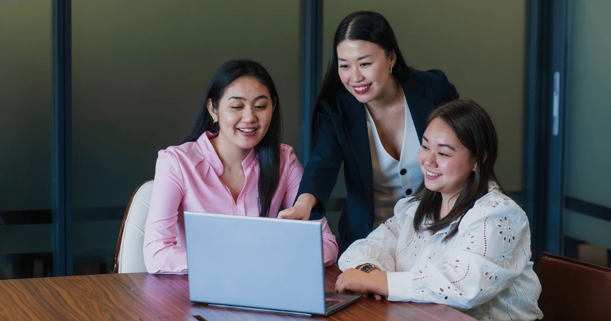 Three women sit around a table, smiling and looking at a laptop screen. One stands behind, pointing at the laptop as they discuss a cost-efficient growth strategy displayed on the screen.