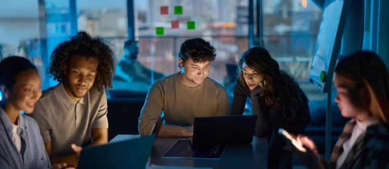 Five young adults sit around a table in a dimly lit modern office, focused on laptops and a smartphone, collaborating as a dedicated offshore team to drive enterprise transformation late into the evening.