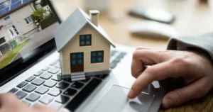 A small model house sits on a laptop keyboard while a person uses the touchpad, with a house listing visible on the screen, symbolizing Real Estate Investment and online home buying.