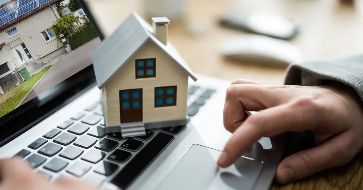 A small model house sits on a laptop keyboard while a person uses the touchpad, with a house listing visible on the screen, symbolizing Real Estate Investment and online home buying.