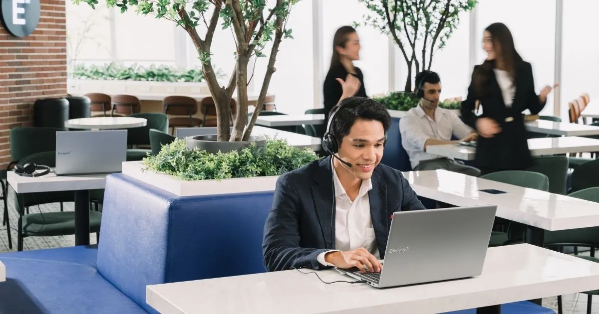 A man wearing a headset sits at a laptop in a modern office lounge with plants and blue seating—a perfect setting for dedicated staffing. In the background, people interact and work on their laptops, supporting back-office scaling.