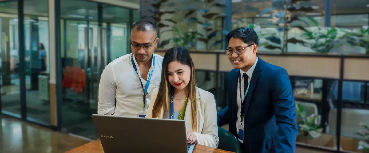 Three colleagues, two men and one woman, look at a laptop screen together and smile in a modern office with glass partitions and plants—a snapshot of teamwork exploring outsourcing as a strategic lifeline for Australia insolvency challenges.