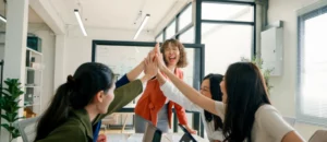 Four women in an office share a group high-five, celebrating in a bright, modern workspace. Their laughter and teamwork reflect strong company culture, making these colleagues a valuable asset to their organization.