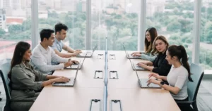 Six people sit across from each other at a long conference table, working on laptops in a modern office with large windows and a city view—discussing cost and how enterprises evaluate workforce models for control & compliance.