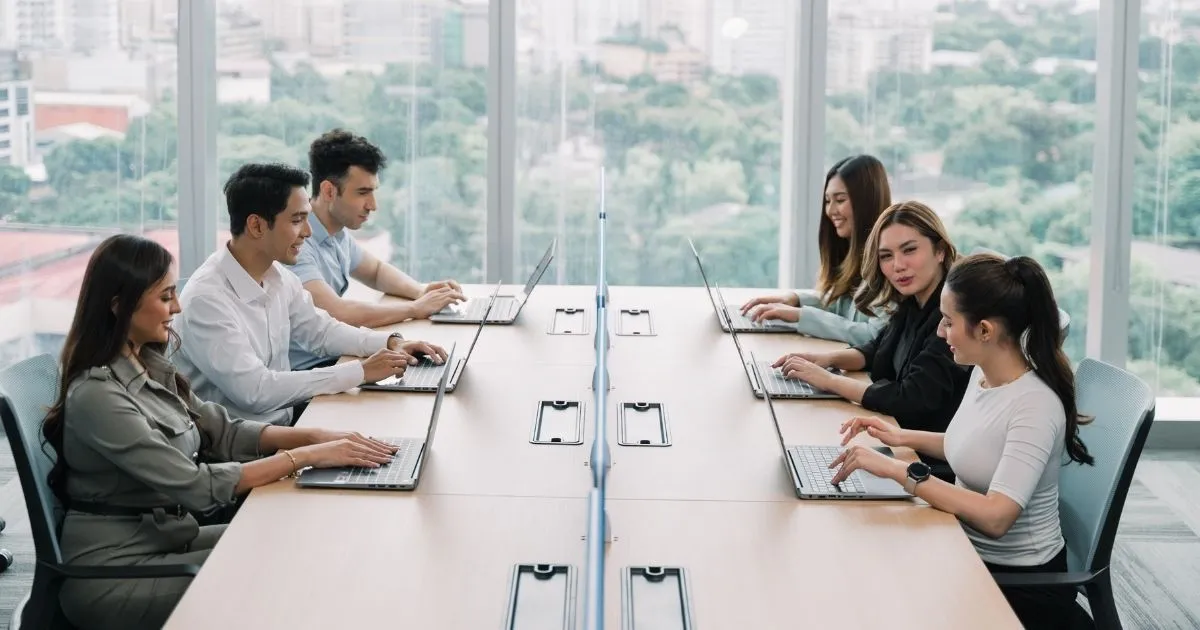 Six people sit across from each other at a long conference table, working on laptops in a modern office with large windows and a city view—discussing cost and how enterprises evaluate workforce models for control & compliance.