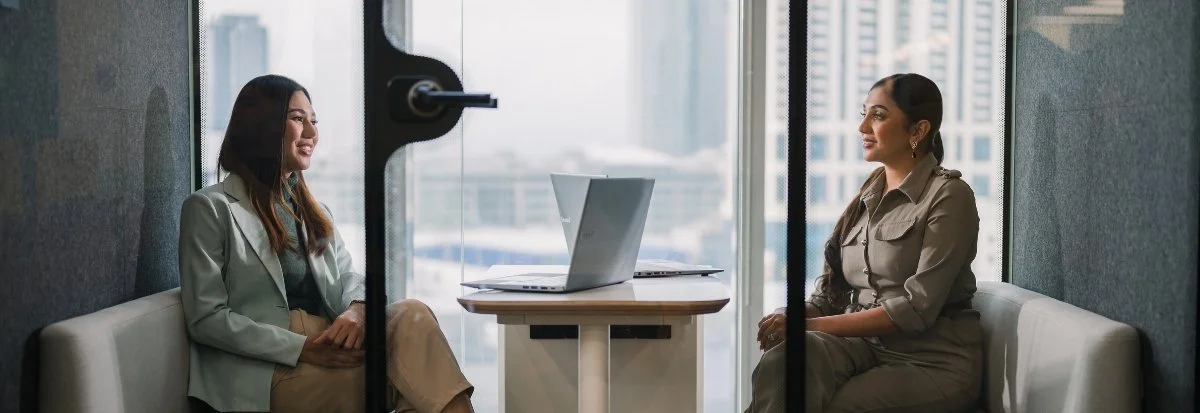 Two women sit across from each other in a modern office meeting room, discussing team scaling. Open laptops rest on the table between them, and large windows reveal a cityscape outside.