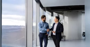 Two businessmen stand near large windows in a modern office, shaking hands and smiling at each other, suggesting a successful meeting or an outsourcing agreement.