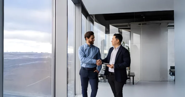 Two businessmen stand near large windows in a modern office, shaking hands and smiling at each other, suggesting a successful meeting or an outsourcing agreement.