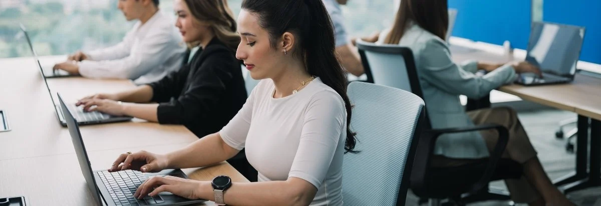 Four people sit in a modern office at desks, working on laptops. The focus is on a woman in a white top typing, with others collaborating in the background—a dynamic scene of team scaling and productivity under large, sunlit windows.