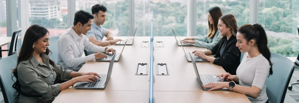 Six people sit across from each other at a long office table, working on laptops. Large windows reveal a cityscape with greenery. The professional, focused atmosphere suggests a team scaling efficiently to handle growing business needs.