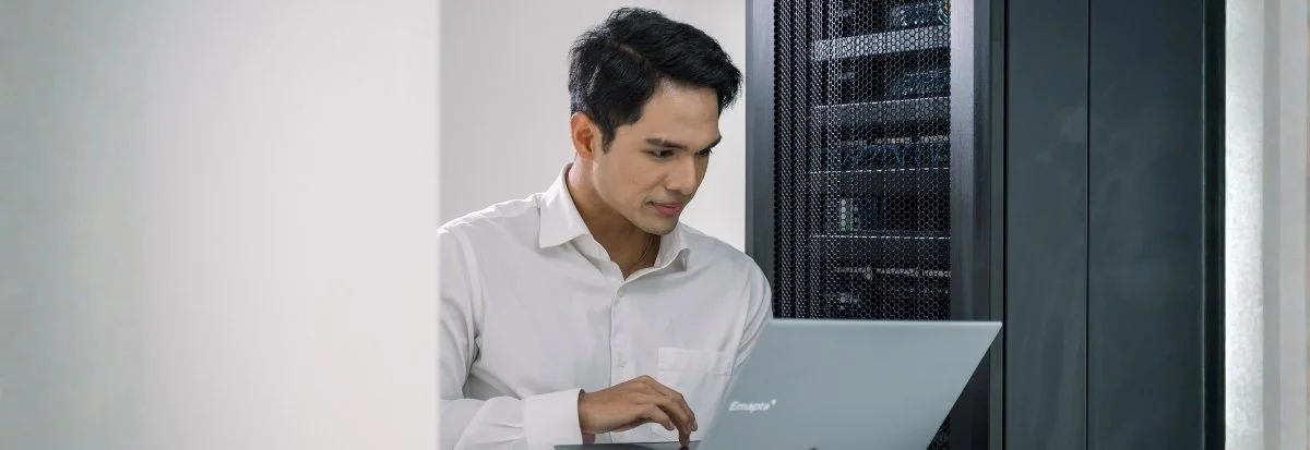 A man in a white shirt is working on a laptop in a server room, standing near a tall server rack, focusing on enterprise scaling and Security.